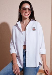 White button-up shirt with rolled sleeves, featuring a stitched teddy bear on the chest. Paired with blue denim and sunglasses.