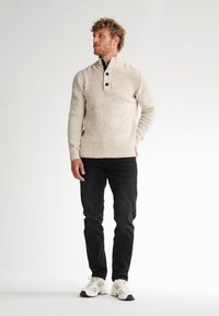 Pullover lavorato a maglia beige chiaro con un motivo testurizzato, colletto con mezza zip e tre bottoni scuri; abbinato a jeans neri e sneakers bianche.