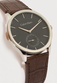 Ρολόι χειρός Calvin Klein με μαύρο καντράν, ασημί κάσα, χρυσούς δείκτες ώρας και δείκτες, μικρό υποκαντράν δευτερολέπτων και καφέ δερμάτινο λουράκι.