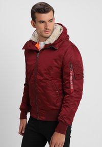 Casaco bomber vermelho com capuz forrado de pelo, punhos canelados, fecho de correr à frente e bolso na manga com texto. Tecido suave com uma textura quente.