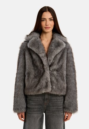 Donna con lunghi capelli scuri, indossa una giacca grigia in pelliccia ecologica e jeans grigio scuro, in piedi davanti a uno sfondo chiaro e semplice.