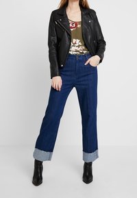 Veste de biker en cuir noir, haut fleuri vert, jean taille haute bleu avec ourlets effilochés et bottines noires.