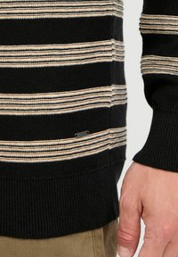 Black knit sweater with cream horizontal stripes, ribbed cuffs and hem, featuring a small logo tag on the lower side.