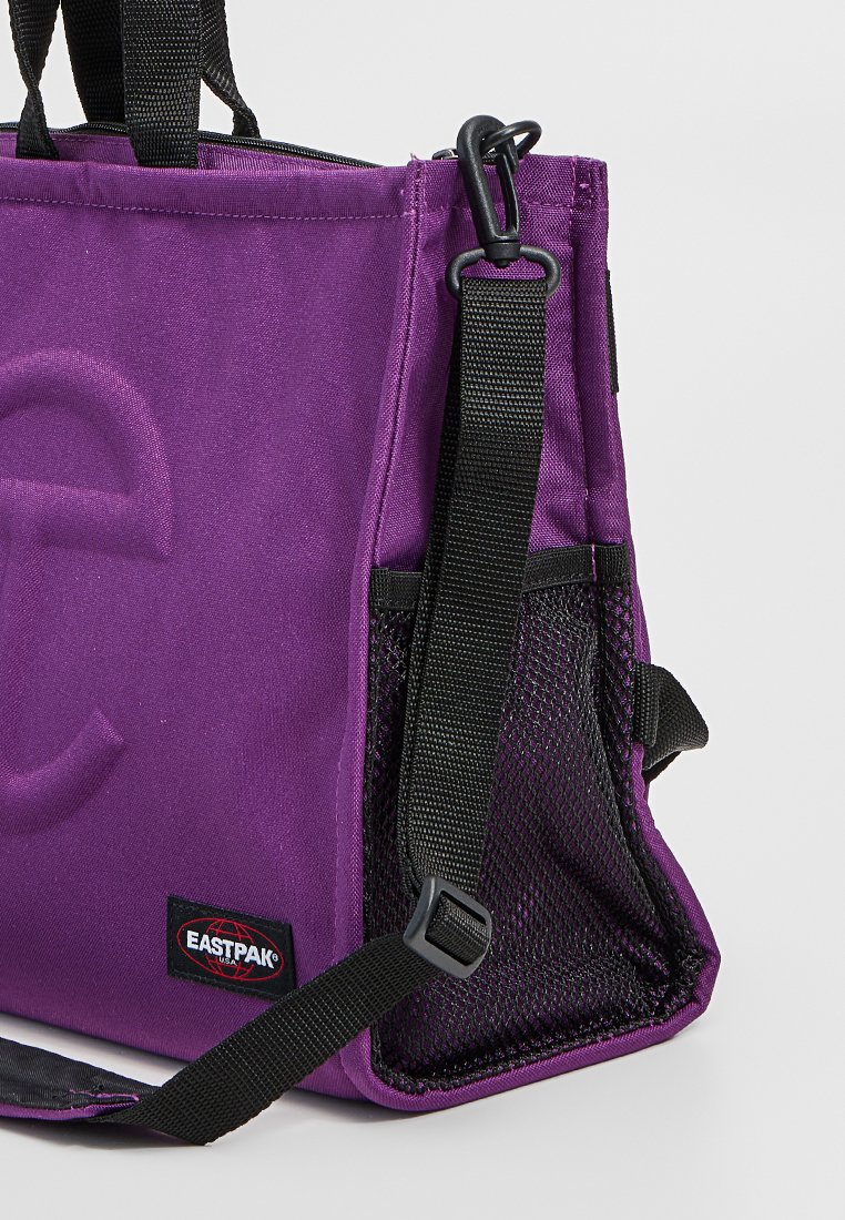 Borsa a tracolla in tela viola con tasca laterale in rete, resistenti manici neri e dettaglio logo in rilievo sulla parte anteriore.