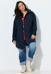Mujer con impermeable azul marino sobre una blusa roja con estampado, vaqueros azules remangados y botas blancas con cordones, de pie contra un fondo blanco.