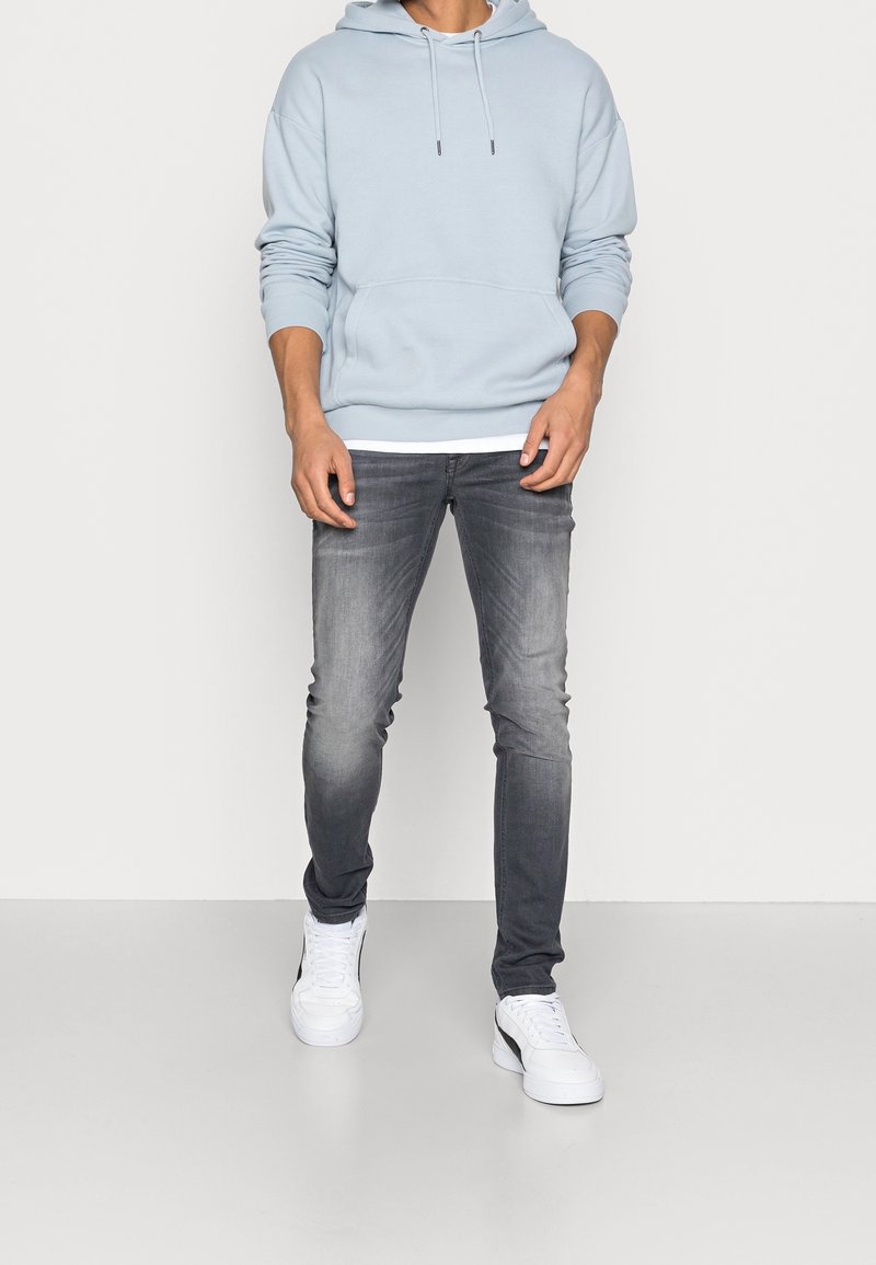 Homme portant un sweat à capuche bleu clair, un jean gris délavé et des baskets blanches, debout devant un fond uni de couleur claire.