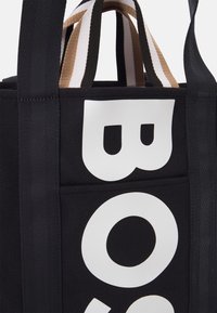 Svart tygväska med en stor vit logotyp. Har robusta svarta och bruna randiga handtag för enkel bärning.