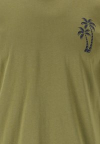 Oliivinvihreä puuvillapaita lyhyillä hihoilla ja tummansininen palmupuu grafiikka vasemmassa rinnassa. Sujuva rakenne, rento malli.