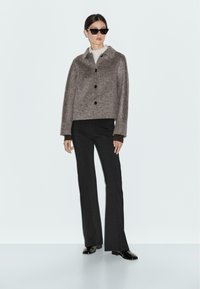 Giacca cropped marrone con colletto, tessuto strutturato e bottoni neri, abbinata a pantaloni neri a zampa d'elefante e occhiali da sole neri.