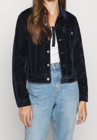 Giacca in corduroy blu navy con un design cropped, bottoni a pressione, due tasche sul petto e maniche lunghe, abbinata a jeans chiari a vita alta.