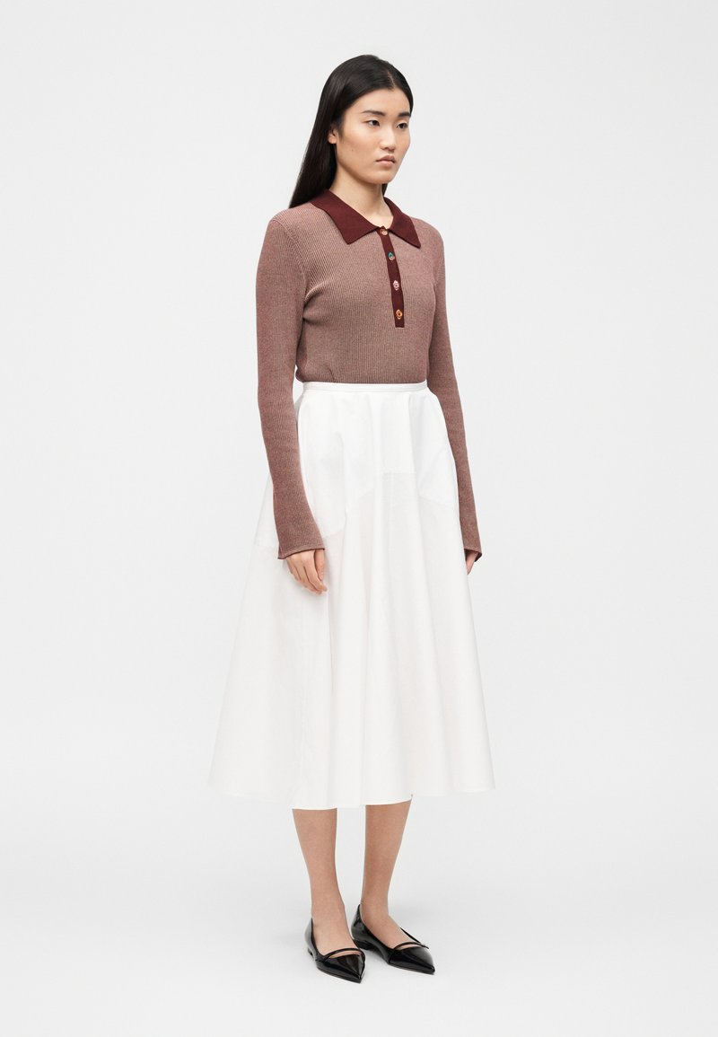 Donna con capelli lunghi e scuri che indossa un top marrone a maniche lunghe a coste con bottoni colorati e colletto, gonna midi bianca e ballerine nere con punta a punta.