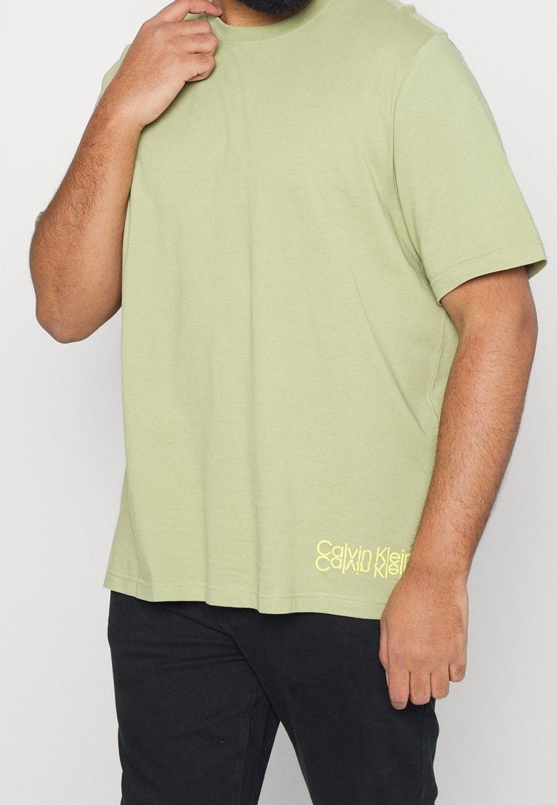 T-shirt vert olive à manches courtes avec un col rond et logo Calvin Klein en jaune sur le bas à gauche. Fabriqué en tissu coton doux.