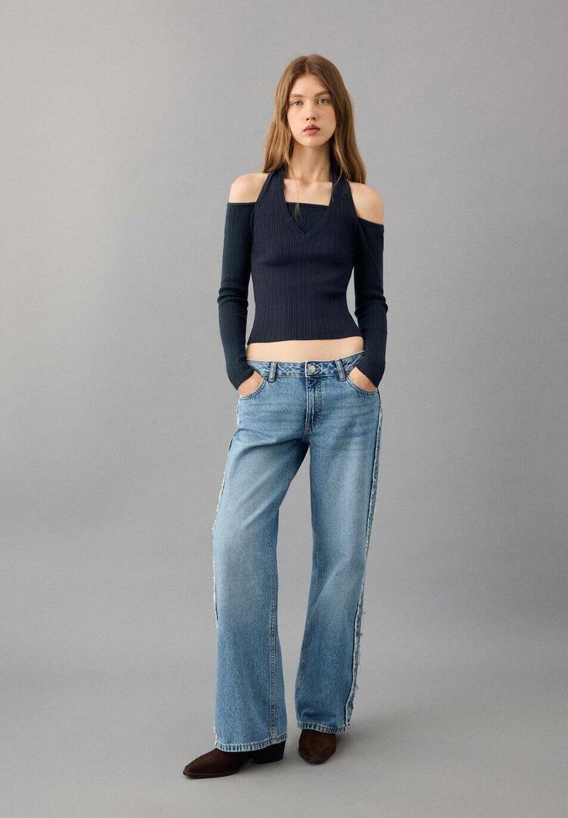 Junge Frau, die ein schwarzes Cold-Shoulder-Halter-Top, lockere hellblaue Jeans und braune Schuhe trägt, vor einem grauen Hintergrund steht.