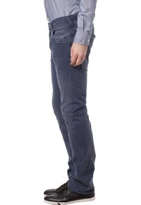 Mężczyzna w niebieskich spodniach z kordurowej tkaniny, w kratkowanej koszuli z długim rękawem i czarnych trampkach, stojący z profilu na białym tle.