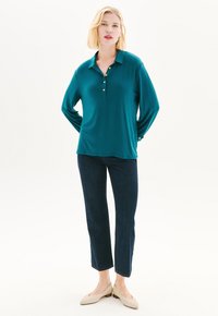 Teal pitkähihaista poolopaitaa, jossa on kolme kultakuppia, yhdistetty tummansinisiin suoraan leikattuihin farkkuihin ja vaaleanbeigeihin matalakorkoisiin kenkiin. Sileä pinta.