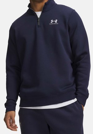 Mann trägt marineblaue Under Armour Viertel-Zip-Sweatshirt und passende Hose, mit weißem Unterhemd sichtbar am Saum.