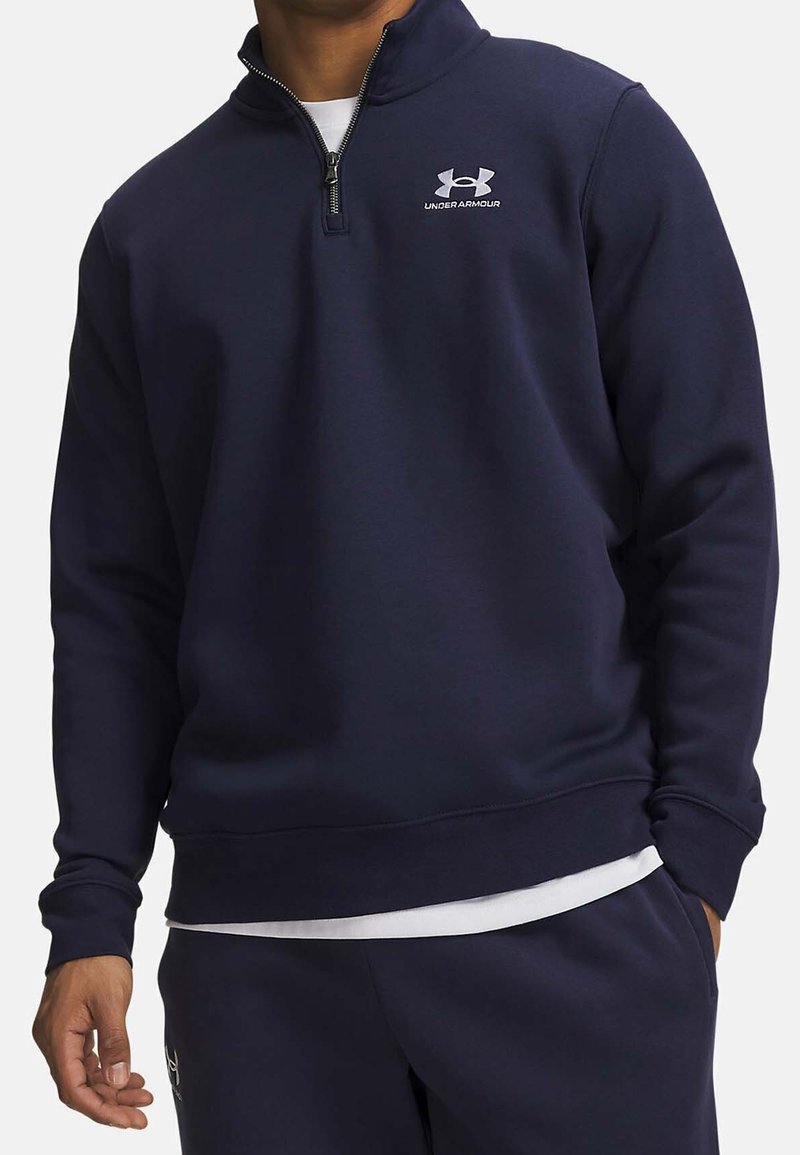 Homme portant un sweat-shirt Under Armour bleu marine à quart de zip et un pantalon assorti, avec un sous-vêtement blanc visible à l'ourlet.