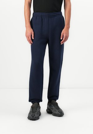 Man wearing navy blue jogger pants, black athletic shoes, and black top, standing against a plain white background.