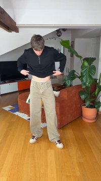Homme en pantalon beige et sweat-shirt noir soulevant légèrement sa chemise dans un salon moderne avec des plantes, un canapé orange, une télévision et un sol en bois.