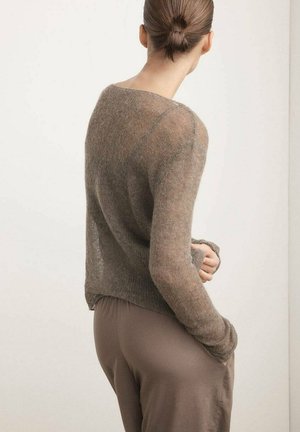 Frau mit hochgesteckten Haaren trägt einen dünnen, braunen Pullover mit langen Ärmeln und lockere taupefarbene Hosen, steht mit einer Hand in der Tasche vor einem einfarbigen Hintergrund.