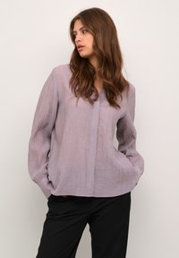 Blusa lunga a maniche con texture a coste verticali in un tenue colore viola, scollatura a V e tasche, abbinata a pantaloni neri. Design casual, vestibilità rilassata.