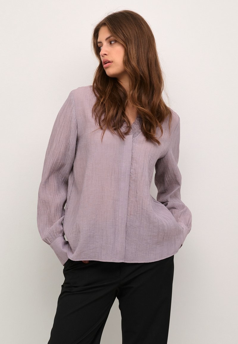 Blusa lunga a maniche con texture a coste verticali in un tenue colore viola, scollatura a V e tasche, abbinata a pantaloni neri. Design casual, vestibilità rilassata.