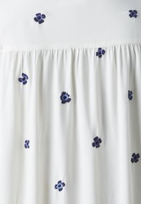 Top bianco in tessuto con texture arricciata, caratterizzato da fiori ricamati blu scuro distribuiti uniformemente sulla parte anteriore per un dettaglio decorativo.