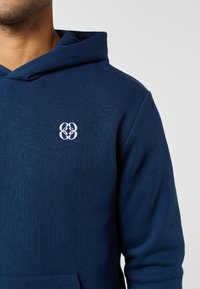 Vyras vilki tamsiai mėlynę su gobtuvu, ant krūtinės yra baltais siūlais siuvinėtas simetriškas logotipas, fonas – šviesiai pilkas ir vientisas.