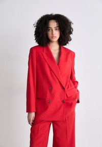 Červený dvouřadý blazer s velkými hnědými knoflíky a odpovídajícími širokými kalhotami, s hlubokým výstřihem do "V" a hladkým povrchem.