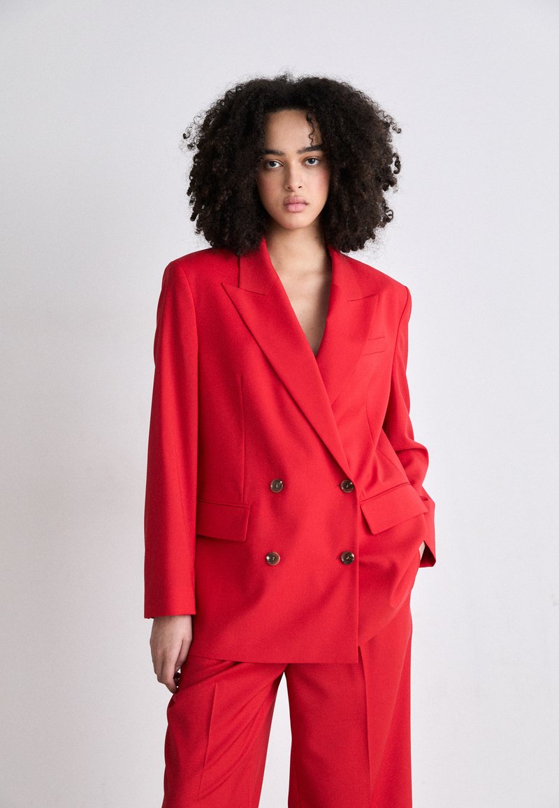 Červený dvouřadý blazer s velkými hnědými knoflíky a odpovídajícími širokými kalhotami, s hlubokým výstřihem do "V" a hladkým povrchem.