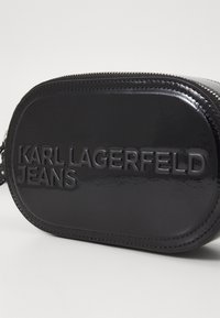 Borsa ovale nera di materiale lucido. Presenta il logo in rilievo "KARL LAGERFELD JEANS" e una chiusura a zip lungo la parte superiore.