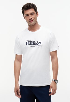 Mann trägt ein weißes Tommy Hilfiger T-Shirt mit dunkelblauem Schriftzug, dunkle Hose und eine silberne Armbanduhr, steht vor einem schlichten weißen Hintergrund.