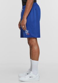 Blå mesh idrettsshorts med elastisk midje, med teksten "NYC 93 KLX" i hvitt og rødt. Brukt med hvite sneakers og sokker.
