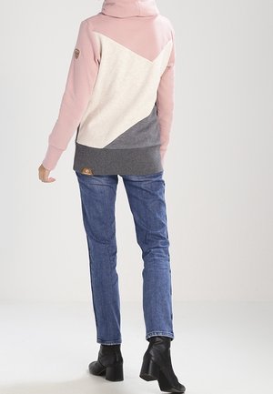 Personne portant un sweat à capuche bicolore rose, crème et gris, un jean bleu et des bottines noires à talons, debout de dos.