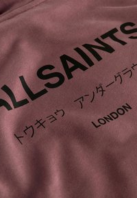 Detailný záber na mauve látku s čiernym textom "ALLSAINTS," japonskými znakmi a nápisom "LONDON" potlačeným na jej povrchu.