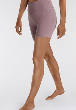 Frau, barfuß gehend, trägt hoch taillierte, mauvefarbene, dehnbare Shorts, dargestellt vom mittleren Oberkörper bis zu den Füßen vor einfachem Hintergrund.