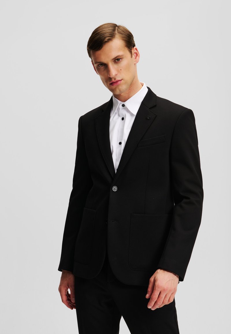 Schwarzer, maßgeschneiderter Blazer mit einem Reverskragen, zwei Vorder- Taschen, einer Knopf- Verschluss und strukturiertem Nähen. Weißes Hemd darunter.
