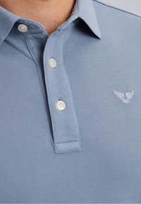 Gros plan sur une personne portant un polo bleu clair avec trois boutons blancs et un petit logo ailé blanc sur la poitrine.