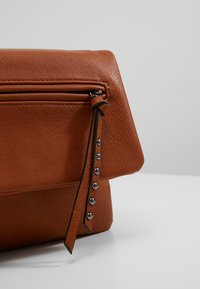 Brun läderklutch med en strukturerad yta, med en dragkedjeficka, ett dekorativt band och metallnitar längs sidan.