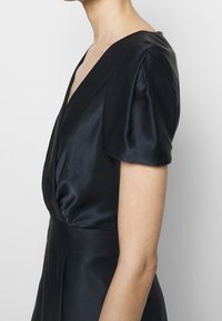 Žena v krátkých rukávech z černého saténového šatu s hlubokým výstřihem do V a přiléhavým pasem, zobrazená z boku na jednobarevném pozadí.