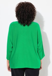 Grünes Strickoberteil mit lockerer Passform, das über drei Viertelärmel und blaue Akzente an den Schultern verfügt. Leichte Textur und glatte Oberfläche.