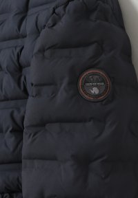 Giubbotto imbottito di color blu navy scuro, con sezioni trapuntate, una texture morbida e una patch rotonda con un logo nei toni del nero e arancione.