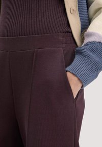 Bordeauxkleurige broeken met een gladde textuur, voorzien van plooien en zijzakken. Gecombineerd met een gemêleerde, geribde gebreide cardigan.