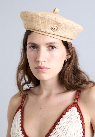 Tkana bež beretka s teksturiranom završnom obradom, s gornjom petljom i izvezenim logotipom. Model nosi halter top od kukičanog materijala s smešnim naglascima.