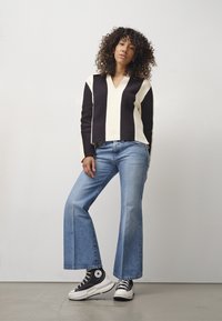 Czarny i kremowy sweter w paski z dekoltem w serek, połączony z jasnoniebieskimi dżinsami w fasonie dzwonów i czarnymi sneakersami za kostkę z białą podeszwą.