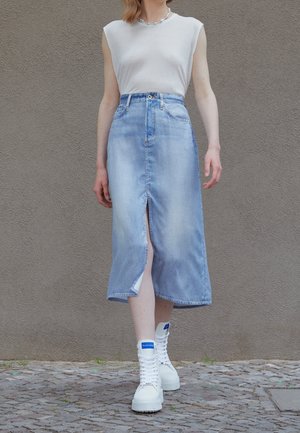 Personne portant un haut blanc sans manches, une jupe midi en denim bleu clair avec une fente à l'avant, des bottes blanches à lacets et un collier de perles.