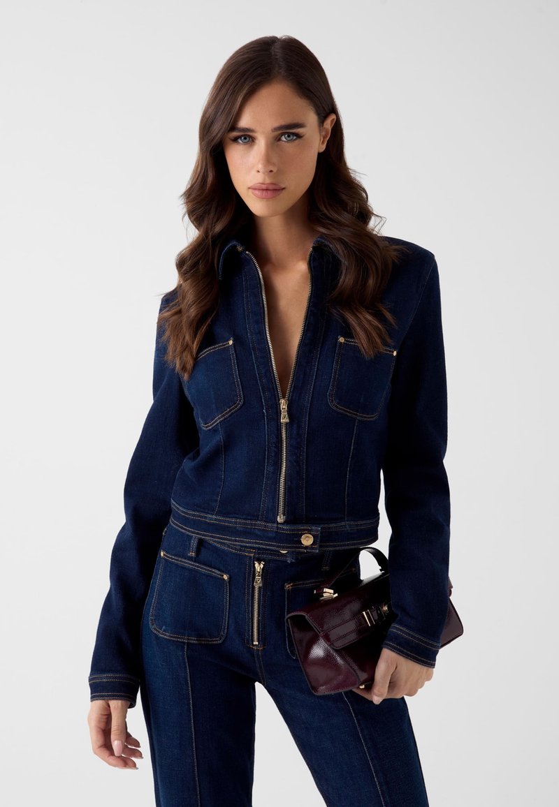 Kombinezon dżinsowy w ciemnym niebieskim kolorze z złotym przeszyciem i suwakami, z dwoma kieszeniami z przodu. Modelka trzyma małą bordową torebkę.