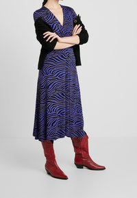 Violetti mekko, jossa mustat seepraraidat ja V-kaula-aukko, yhdessä mustan neulotun neuletakin kanssa. Yhdistetty punaisiin länkkärityylisiin nilkkureihin.