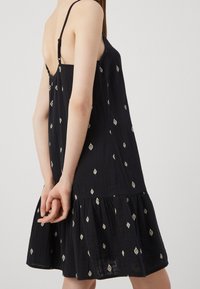 Musta hihaton mekko, jossa on löysä istuvuus. Mekossa on valkoisia timantinmuotoisia kuvioita, röyhelöhelma ja säädettävät hihnat.