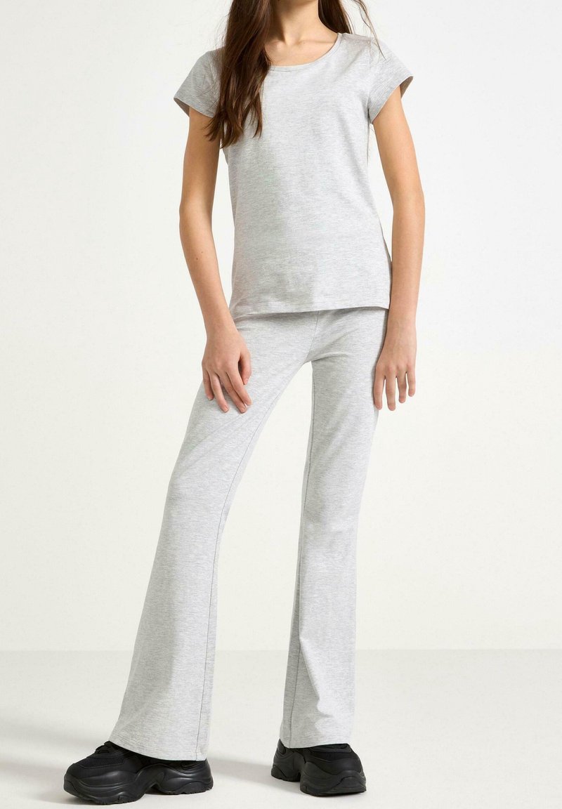 Jeune femme portant un haut à manches courtes gris clair et un pantalon évasé assorti, debout sur fond blanc avec des chaussures épaisses noires.